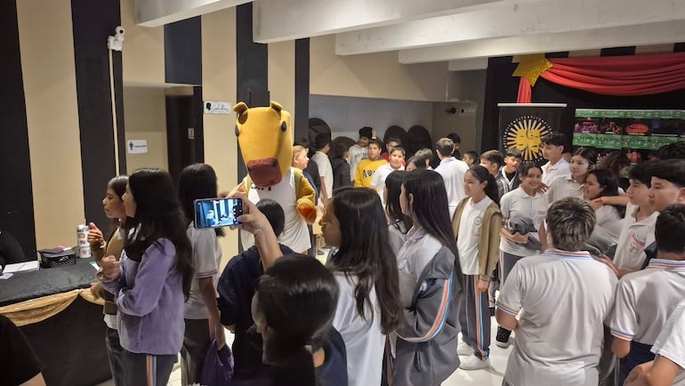 Grupo de jóvenes con camisetas blancas, observando atento a un personaje disfrazado de carpincho; ambiente festivo y de curiosidad.