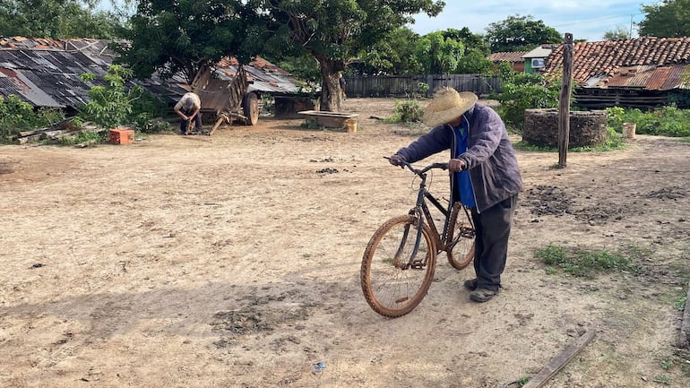 Hombre mayor con sombrero y chaqueta gris sostiene una bicicleta, mientras otro trabaja agachado en tierra suelta.