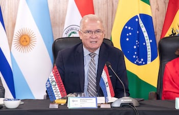 Dr. Emiliano Rolón Fernández habla en un panel, con traje oscuro y gafas, junto a banderas y una mujer escuchando atentamente.
