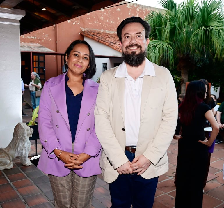 Mujer de cabello corto y abrigo morado sonríe, junto a un hombre de barba y abrigo claro, en un evento social al aire libre.