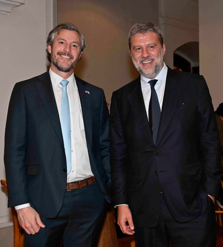Dos hombres sonrientes, uno en traje oscuro y corbata clara, otro en traje negro y corbata oscura, posando en un evento formal.