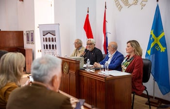 Hombre con cabello gris habla en mesa con otros dos hombres y una mujer, rodeados de asistentes y banderas en el fondo.
