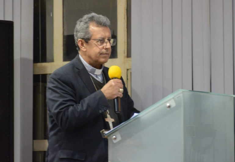 Hombre canoso en camisa clara y saco oscuro habla con micrófono. Ambiente de auditorio con paredes y podio frente a él.