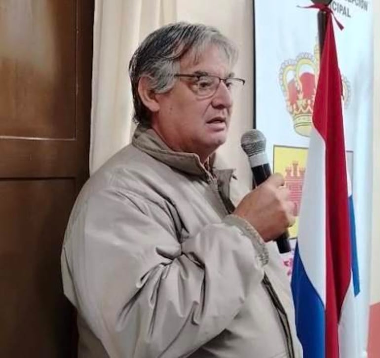Hombre canoso con gafas y abrigo claro, sosteniendo un micrófono frente a un mural con la bandera paraguaya.