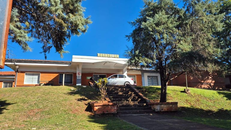 Edificio de urgencias con letrero visible, césped verde y árbol alrededor, vehículo estacionado frente a la entrada.