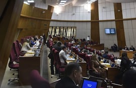 Sala de deliberaciones con aproximadamente 20 personas, algunos sentados en escritorios, ambiente formal y profesional.