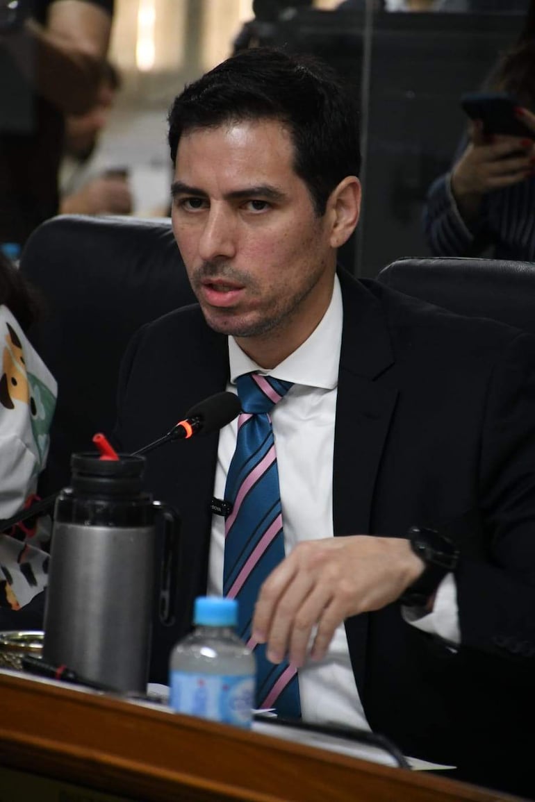 Alvaro Grau sentado en mesa con traje negro y corbata azul y rosa, conversando con persona a su izquierda en entorno formal.