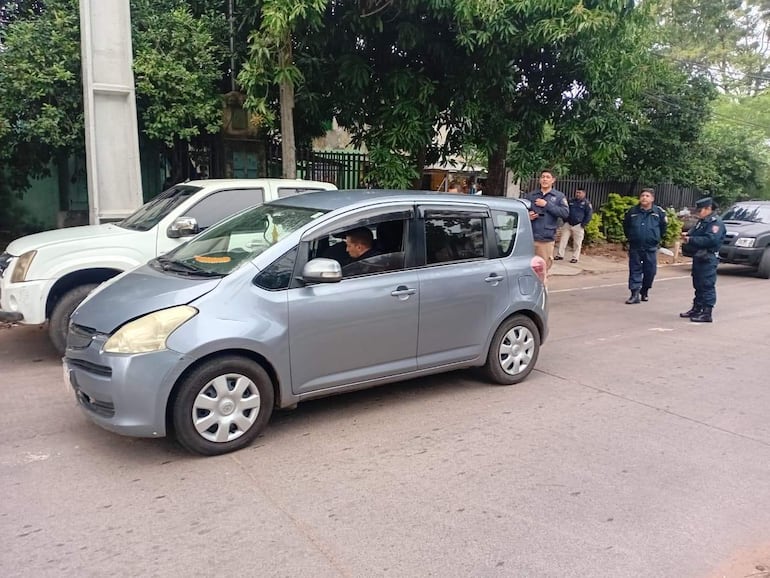 Vehículo gris con conductor hablando mientras policías en uniforme observan en la acera, rodeados de vegetación.