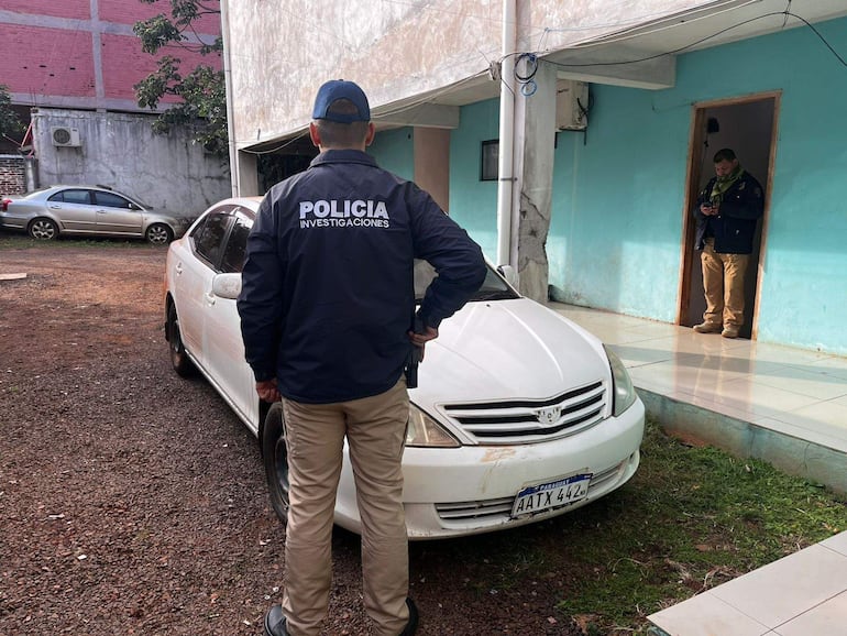 Oficial de policía en abrigo con inscripción, vigilando mientras otro hombre utiliza un dispositivo móvil en un ambiente residencial.