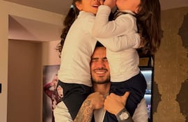 Antonio Sanabria con tatuajes sonríe mientras sostiene a sus hijas en hombros, todas vestidas con camisetas blancas.