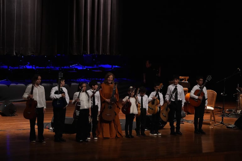 Ocho personas, incluyendo una mujer en vestido naranja y seis niños en camisas blancas, tocan guitarras en un auditorio iluminado.