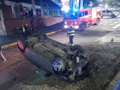Dos agentes de seguridad con chalecos en la escena de un auto volcado, bomberos con luces intermitentes en el fondo.