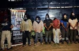 Siete hombres esposados con capuchas negras y un agente de policía uniformado, en un ambiente oscuro de operativo policial.