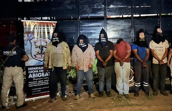 Siete hombres esposados con capuchas negras y un agente de policía uniformado, en un ambiente oscuro de operativo policial.