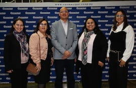 Cinco adultos bien vestidos sonríen en un evento, destacando a un hombre en chaleco gris y mujeres con bufandas coloridas.