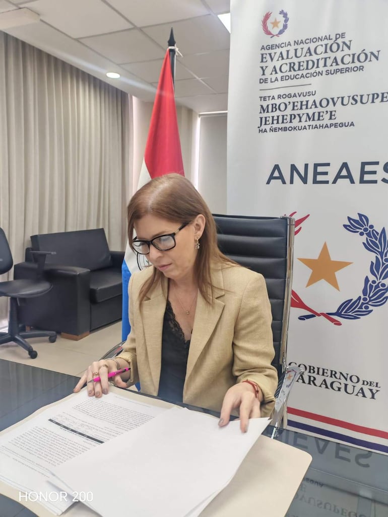 Adriana Pesoa sentada en una oficina, concentrada en documentos, con una bandera de Paraguay a su izquierda.