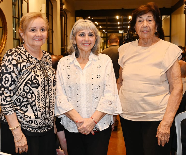 Tres mujeres sonrientes: una con blusa blanca bordada, otra en patrón blanco y negro, y la última en blusa clara, en ambiente cálido.