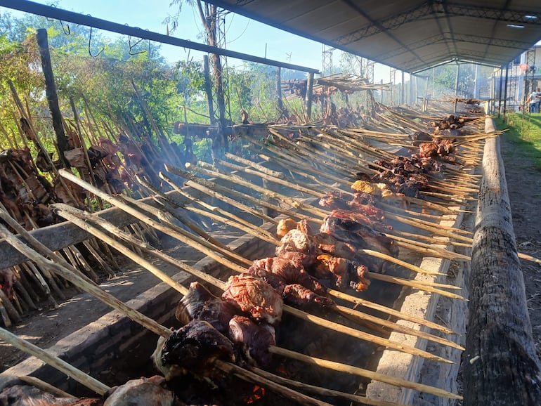 Parrilla al aire libre con varias piezas de carne asándose y humo visible en un ambiente festivo.