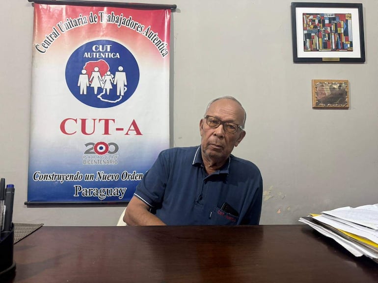 Hombre mayor con gafas y camiseta azul sentado en una mesa con documentos, detrás un cartel de CUT Auténtica en ambiente de oficina.