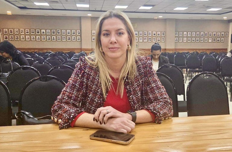 Mujer de cabello rubio con blazer colorido y blusa roja, sentada en una mesa, sosteniendo un teléfono móvil.