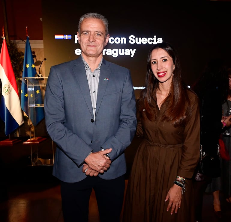 Un hombre canoso con traje gris sonríe junto a una mujer de vestido marrón en un ambiente elegante con banderas de Suecia y Paraguay.
