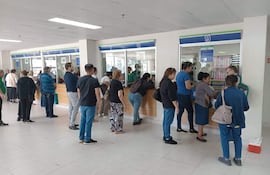 Grupo de personas esperando en mostrador, algunos con bolsas, en un ambiente iluminado y organizado.