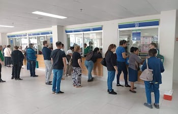 Grupo de personas esperando en mostrador, algunos con bolsas, en un ambiente iluminado y organizado.