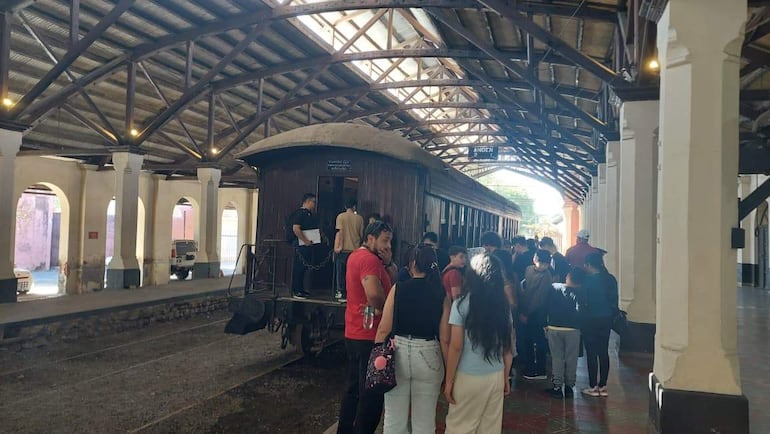 Grupo de 20 personas en el andén de una estación, observando un tren de madera antigua, algunas vistiendo vaqueros, con expresiones de atención.