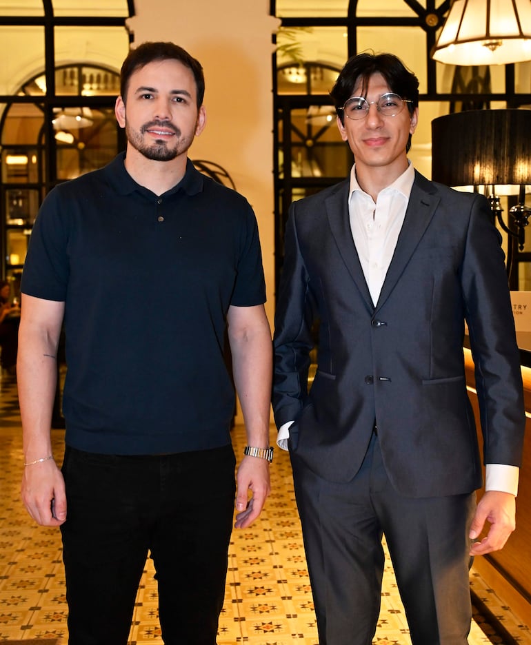 Dos hombres, uno con camiseta negra y otro con traje oscuro y camisa blanca, sonríen en un elegante hotel.