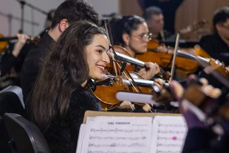 Joven violinista sonriente en el centro, rodeada de músicos en trajes negros tocando en un auditorio iluminado.