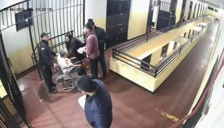 Persona mayor en silla de ruedas con camisa blanca, rodeada de una mujer de cabello oscuro y un guardia con vestimenta oscura en un pasillo amarillo.