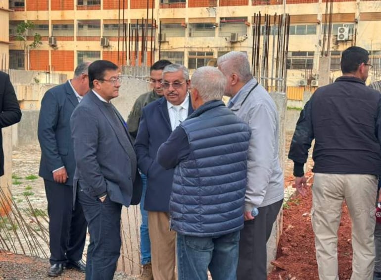 Seis personas discuten en un sitio de construcción; uno con saco oscuro y corbata, otro con chaleco azul, frente a una edificación incompleta.
