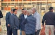 Seis personas discuten en un sitio de construcción; uno con saco oscuro y corbata, otro con chaleco azul, frente a una edificación incompleta.