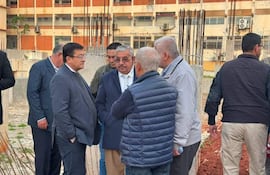 Seis personas discuten en un sitio de construcción; uno con saco oscuro y corbata, otro con chaleco azul, frente a una edificación incompleta.