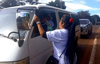Mujer con camiseta blanca y lazo rojo sostiene un recipiente de colecta cerca de una furgoneta blanca. Hombre al volante observa.