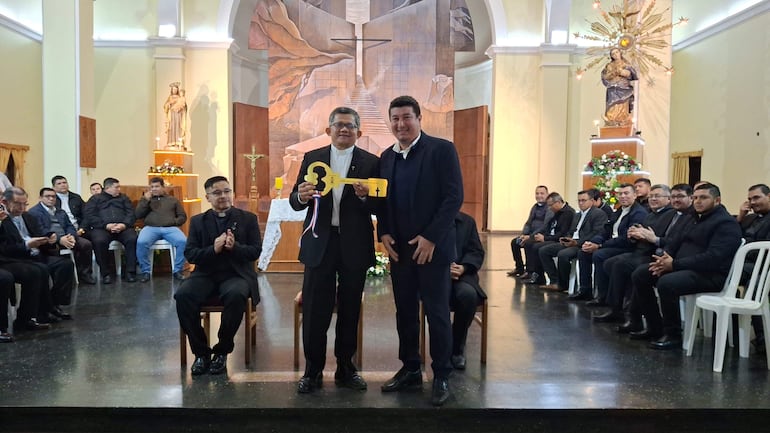 Dos hombres en traje, uno sostiene una llave amarilla, rodeados de público vestido de negro en un ambiente solemne.
