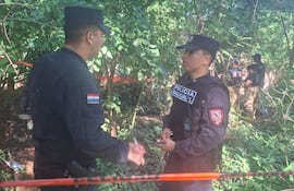 Dos policías en uniforme negro conversan en el bosque, con otros uniformados trabajando en el fondo y cinta de precaución visible.