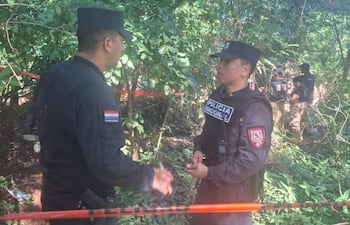 Dos policías en uniforme negro conversan en el bosque, con otros uniformados trabajando en el fondo y cinta de precaución visible.