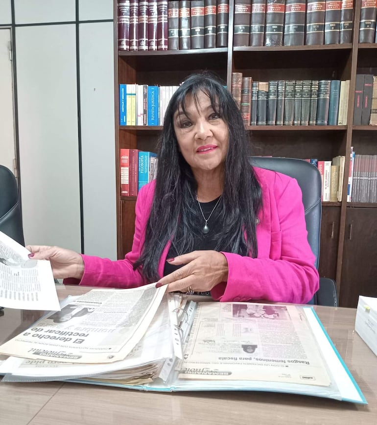Mujer con chaqueta rosa y cabello oscuro, mirando a la cámara mientras revisa documentos en su oficina.