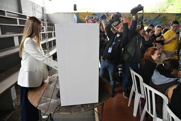 Soledad Núñez con blazer blanco, interactuando con una urna electoral mientras fotógrafas la capturan en un entorno lleno de público sonriente.