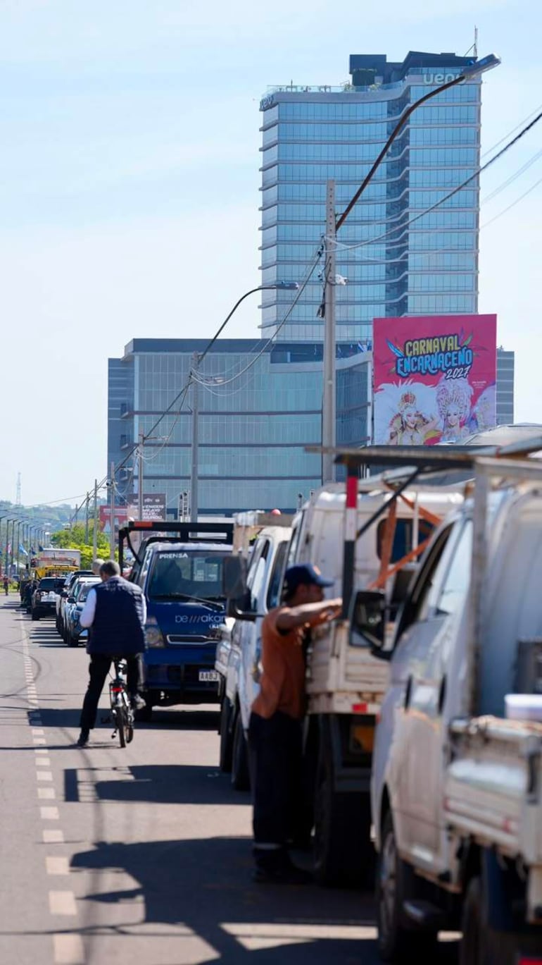 Ciclista avanza por la calle junto a un hombre junto a un vehículo, con un edificio moderno y cartel del Carnaval Encarnaceno al fondo.