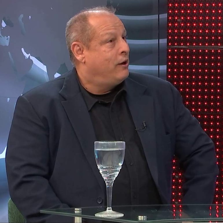 Carlos Díaz Casabianca, con cabello canoso y camisa negra, habla en un set iluminado con luces rojas, frente a una mesa de vidrio.