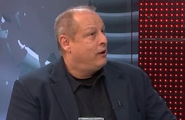 Carlos Díaz Casabianca, con cabello canoso y camisa negra, habla en un set iluminado con luces rojas, frente a una mesa de vidrio.