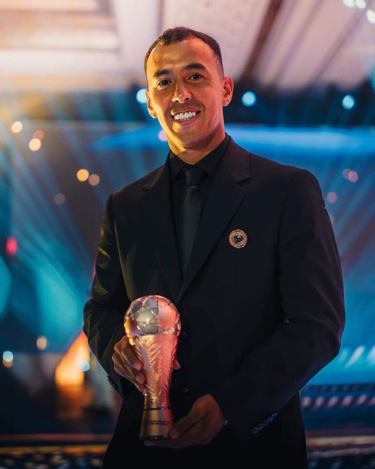 Alejandro Romero Gamarra Kaku sonríe con un trofeo en mano, vistiendo un traje negro en un ambiente festivo y formal.