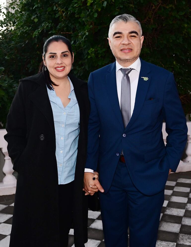 Jeison Granados sonriente en esmoquin azul oscuro, junto a mujer con abrigo negro y camisa de rayas, en un jardín.