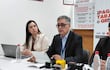 Báez en traje oscuro habla en mesa con laptop, acompañado de una mujer con abrigo claro y un hombre en segundo plano.