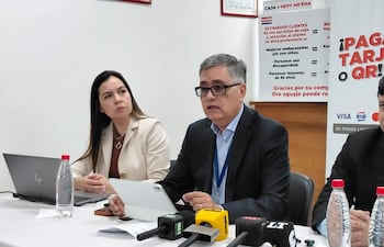 Báez en traje oscuro habla en mesa con laptop, acompañado de una mujer con abrigo claro y un hombre en segundo plano.