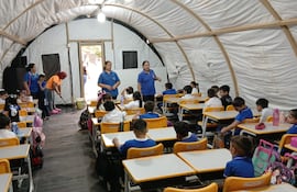 Tres profesoras dando clases bajo una carpa de campamento. Mientras tanto el personal de aseo seca el agua que filtra del compresor del acondicionador de aire.