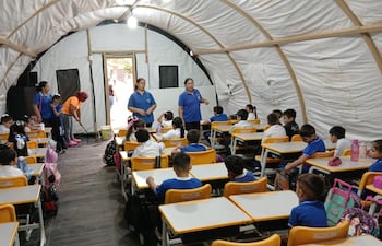 Tres profesoras dando clases bajo una carpa de campamento. Mientras tanto el personal de aseo seca el agua que filtra del compresor del acondicionador de aire.