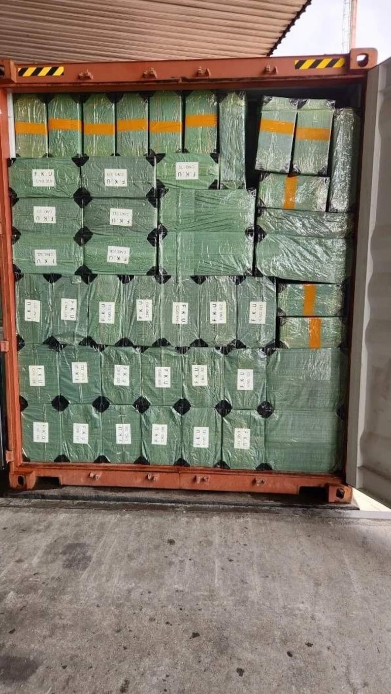Cajas apiladas en un contenedor en puerto. Etiquetas blancas visibles, sin presencia de personas en la imagen.